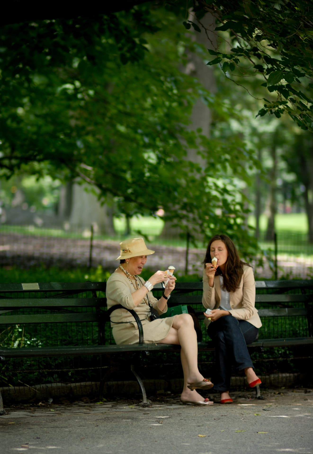 Two women sharing ice cream on a park bench in dappled shade